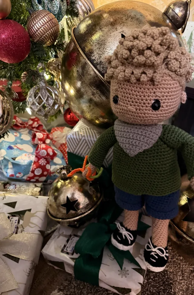 A crocheted boy doll with curly hair, a green sweater with a grey bandana, blue shorts and laced sneakers, standing among Christmas presents