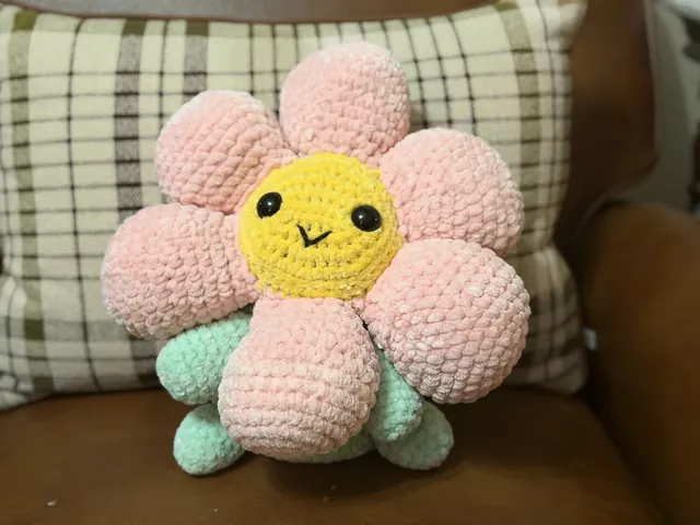 A large plush crocheted flower with blush-pink petals, a smiling yellow face and mint-green leaves