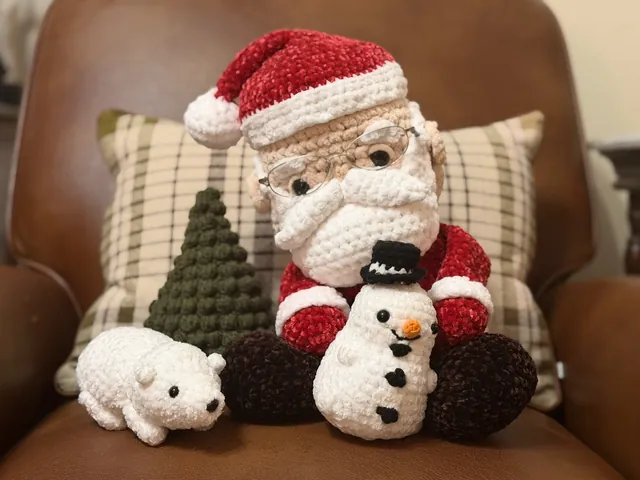 A plush red velvet Santa wearing real glasses, holding a crocheted snowman, beside a polar bear and a green bobble-stitch Christmas tree