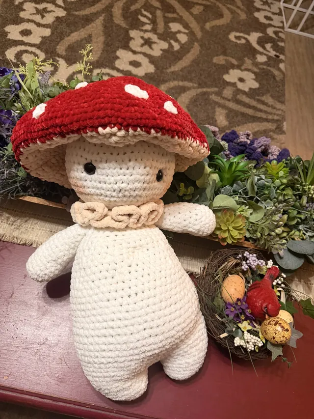 A cream crocheted figure wearing a red toadstool cap with white spots and a ruffled collar