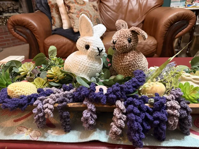 A white and a brown crocheted velvet bunny sitting among crocheted purple lavender sprigs and pastel eggs