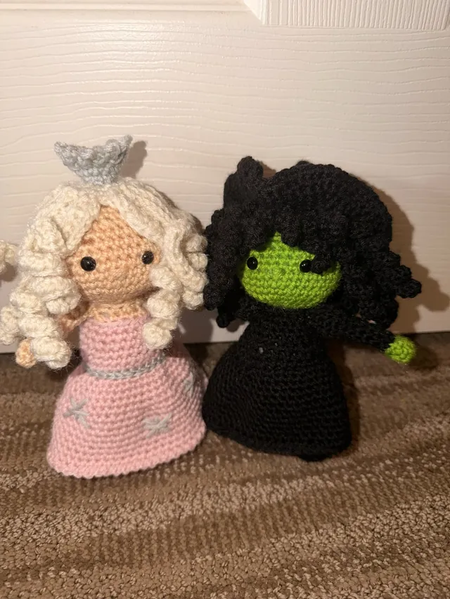 Two crocheted witch dolls: one with blonde curls, a silver crown and a pink gown, the other green-skinned with black curls and a black dress